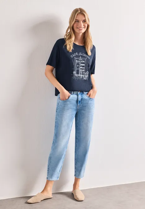 T-Shirt mit Rundhals und Frontprint darkest blue Frau in navy Shirt mit Print, hellblauen Jeans und beigen Slippern steht vor weißer Wand, Hände in den Taschen.