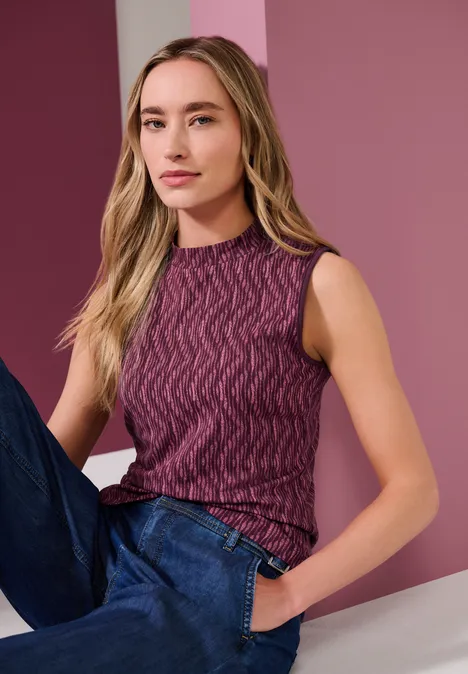 Jeune femme en pull sans manches violet et jean, appuyée devant un mur rose.