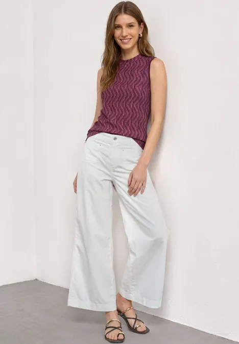 Femme en débardeur violet et pantalon blanc ample, debout dans un coin, portant des sandales.