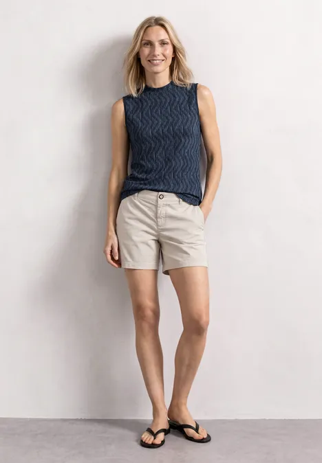 Frau in blauem Top und beigen Shorts mit Flip-Flops, vor hellem Hintergrund, lächelt entspannt.