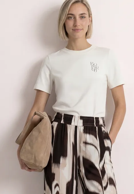 T-Shirt mit Rundhals und Chestprint off white Frau im weißen T-Shirt mit weit geschnittener schwarz-weißer Hose hält eine beige Veloursledertasche.