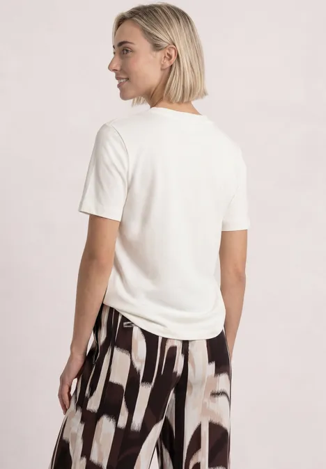 T-Shirt mit Rundhals und Chestprint off white Frau mit kurzem blondem Haar trägt weißes T-Shirt und gemusterte, weite Hose, Rückenansicht.