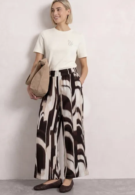 T-Shirt mit Rundhals und Chestprint off white Frau in cremefarbenem T-Shirt und weiten, schwarz-weißen Hosen mit abstraktem Muster, Beuteltasche.