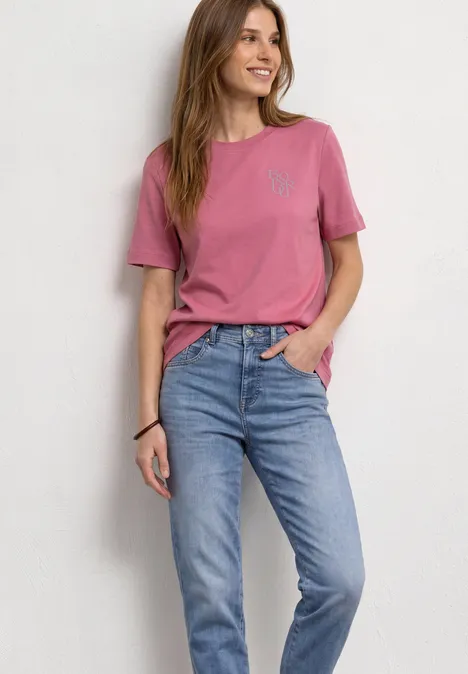 T-Shirt mit Rundhals und Chestprint blush berry Frau in rosa T-Shirt und blauen Jeans steht lächelnd vor weißem Hintergrund.