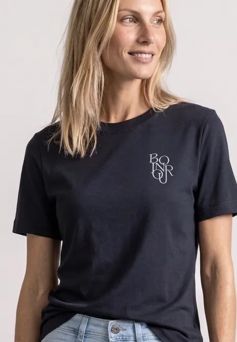 Eine lächelnde Frau in schlichtem schwarzen T-Shirt mit kleinem weißen Logo auf der Brust.