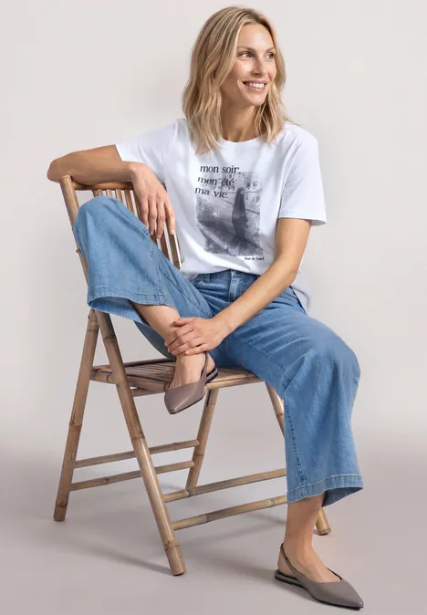 T-Shirt mit Rundhals und Fotoprint White Eine Frau sitzt entspannt seitlich auf einer Holzleiter, trägt weißes T-Shirt, weite Jeans und spitze, taupefarbene Schuhe.