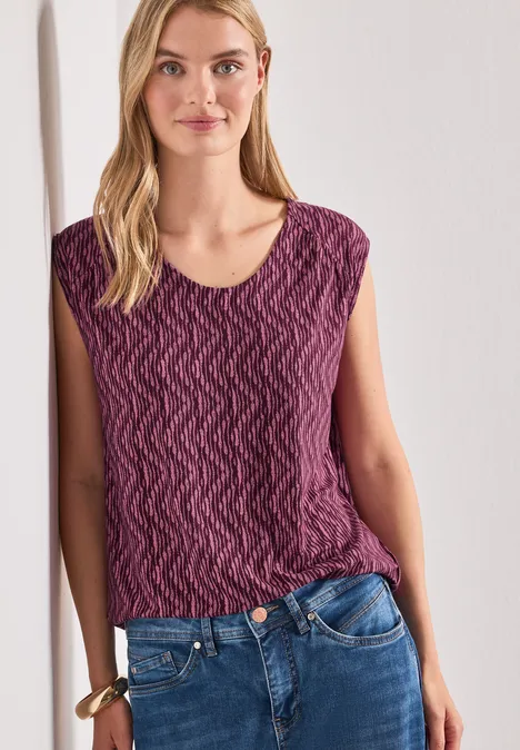 Femme penchée contre un mur blanc, portant un top violet texturé et un jean