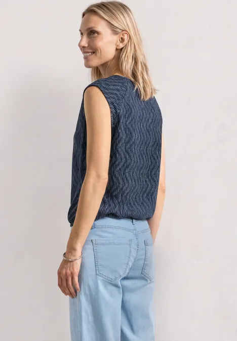 Vrouw van achteren in een mouwloze, golvende navyblauwe top en lichtblauwe spijkerbroek voor een neutrale achtergrond.
