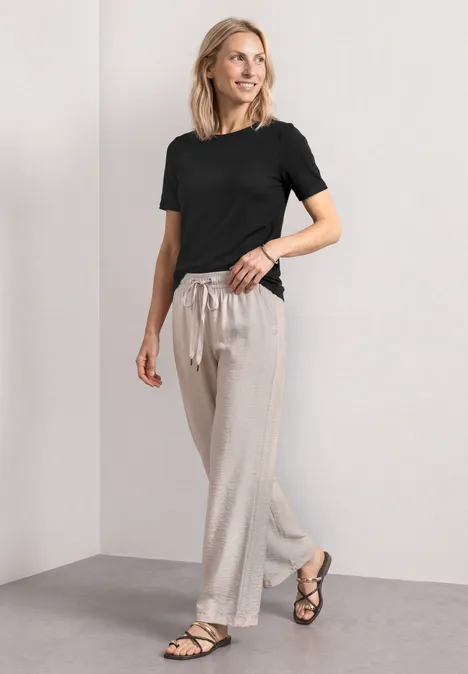 Basic Shirt met ronde hals in Unifarbe Black Vrouw in een minimalistische setting met zwart T-shirt, lichte linnen broek met trekkoord en zwarte sandalen.