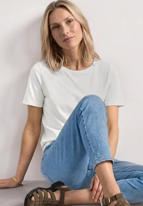 Frau sitzt entspannt an einer Wand in weißem T-Shirt und blauen Jeans, ein Bein angewinkelt.