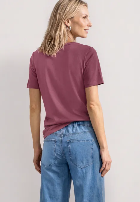 Frau von hinten in lila T-Shirt und blauer Jeans vor neutralem Hintergrund.