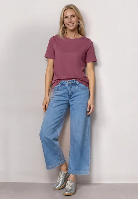 Frau steht vor neutralem Hintergrund in rosa T-Shirt, blauen Marlene-Jeans und silbernen Sneakern.
