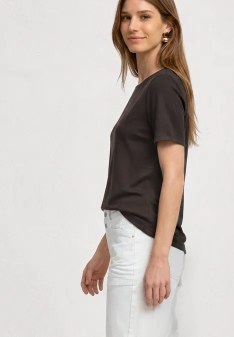 Seitliches Profilfoto einer Frau in grauem T-Shirt und hellen Jeans vor weißem Hintergrund.