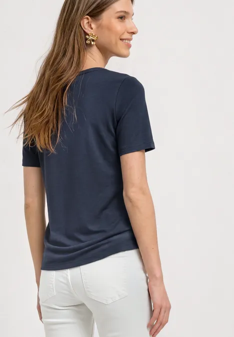 Eine Frau steht seitlich, trägt ein dunkelblaues T-Shirt und weiße Hose, lächelt.