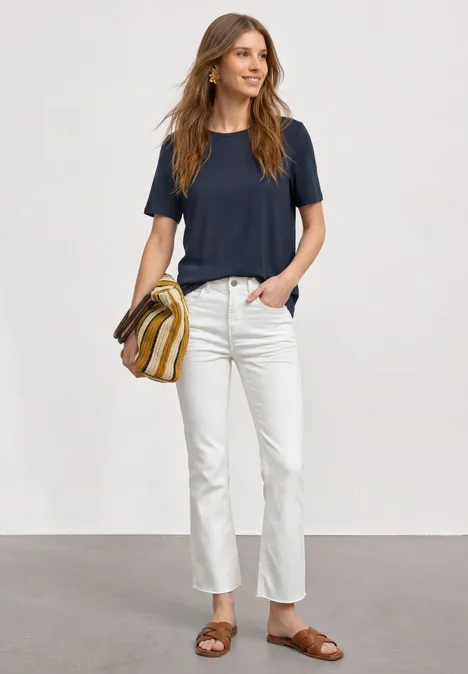 Frau in dunkelblauem T-Shirt, weißen Jeans, braunen Sandalen und gestreifter Tasche vor grauem Hintergrund.