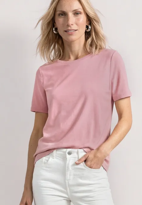 Eine Frau in rosa T-Shirt und weißen Jeans steht lächelnd vor hellem Hintergrund.