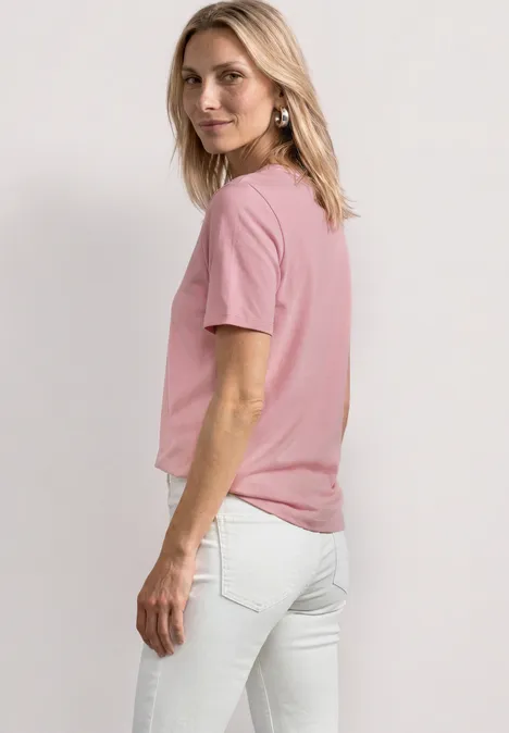 Seitliche Ansicht einer Frau in rosa T-Shirt und weißen Jeans vor hellem Hintergrund.