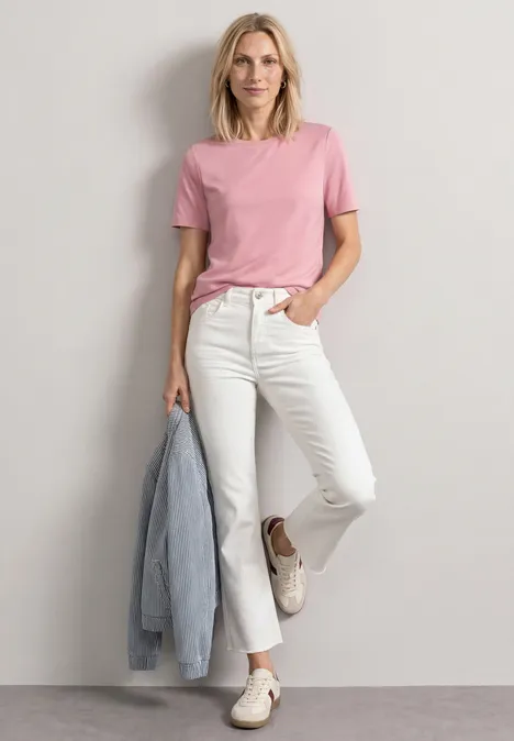 Frau in rosa T-Shirt und weißen Jeans lehnt lässig an der Wand, hält eine Jeansjacke in der Hand.