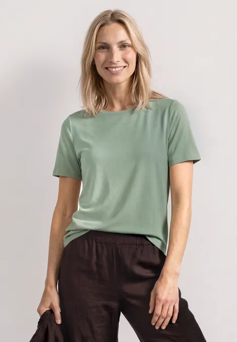 Frau trägt schlichtes mintgrünes T-Shirt und braune Leinenhose vor hellem, schlichten Hintergrund.