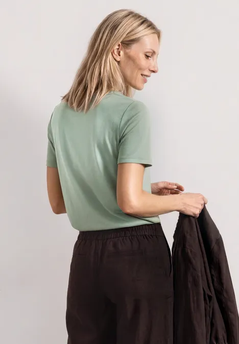 Frau mit blonden Haaren von hinten in mintgrünem T-Shirt, hält eine schwarze Jacke.