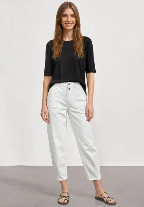 Basic Shirt met ronde hals in Unifarbe Black Vrouw in een witte jeans, zwart T-shirt en sandalen tegen een neutrale achtergrond.
