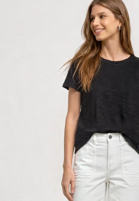 Basic Shirt met ronde hals in Unifarbe Black Een vrouw in een zwart t-shirt en witte broek voor een lichte achtergrond.
