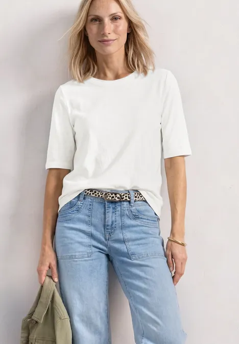Frau im weißen T-Shirt, halb in hellblaue Jeans mit Leopardenmuster-Gürtel gesteckt, hält olivgrüne Jacke.