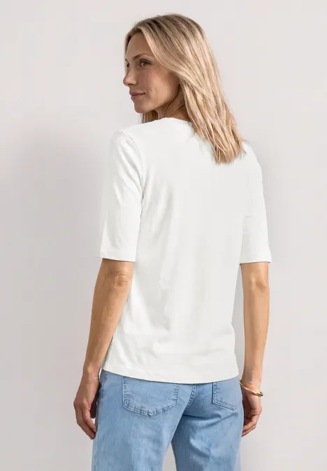 Rückansicht einer Frau mit schulterlangem blondem Haar, weißem Halbarmshirt und blauen Jeans.