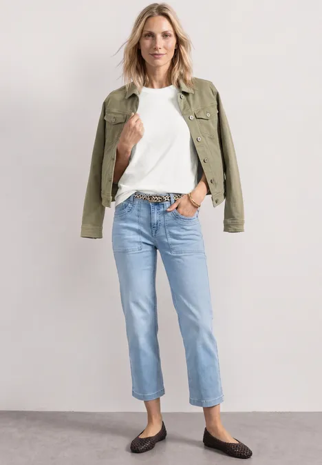 Frau in weißem T-Shirt, Jeans, grüner Jacke und flachen schwarzen Schuhen.