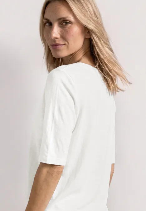 Seitlich abgebildete Person mit halblangem blondem Haar trägt ein schlichtes weißes T-Shirt.