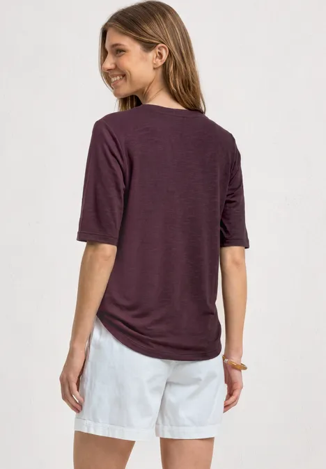 Basic Shirt met ronde hals in Unifarbe rustic rouge Vrouw van achteren met een losvallend donkerpaars shirt en witte korte broek voor een lichte achtergrond.