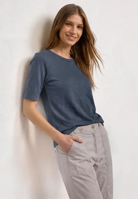 Basic Shirt met ronde hals in Unifarbe ashen blue Vrouw leunt tegen een lichte muur, draagt een donkerblauw T-shirt en lichtbeige broek, hand in broekzak.