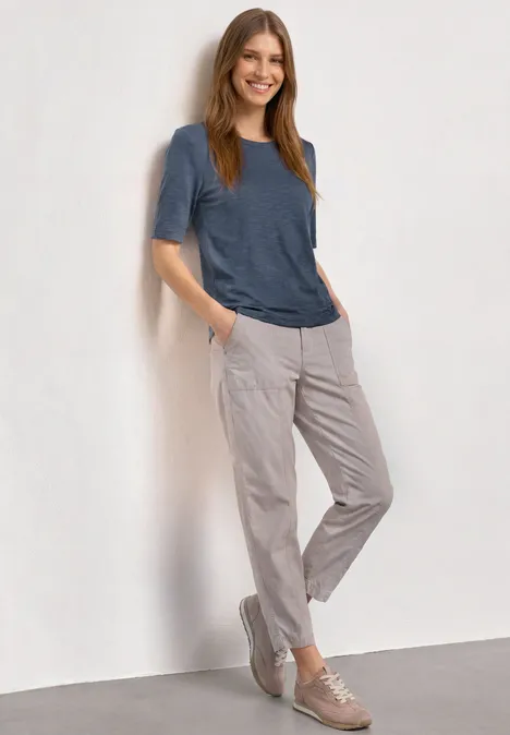 Basic Shirt met ronde hals in Unifarbe ashen blue Vrouw leunt nonchalant tegen een muur, draagt een blauw T-shirt, lichtgrijze broek en beige sneakers.