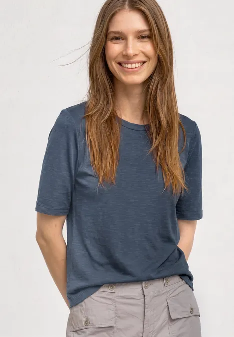 Basic Shirt met ronde hals in Unifarbe ashen blue Vrouw draagt blauw T-shirt en grijze broek, glimlacht voor witte achtergrond.