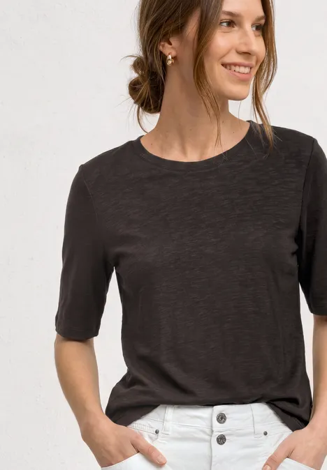 Basic Shirt met ronde hals in Unifarbe black coffee Vrouw in donker T-shirt en witte broek met knopen, handen in de zakken.
