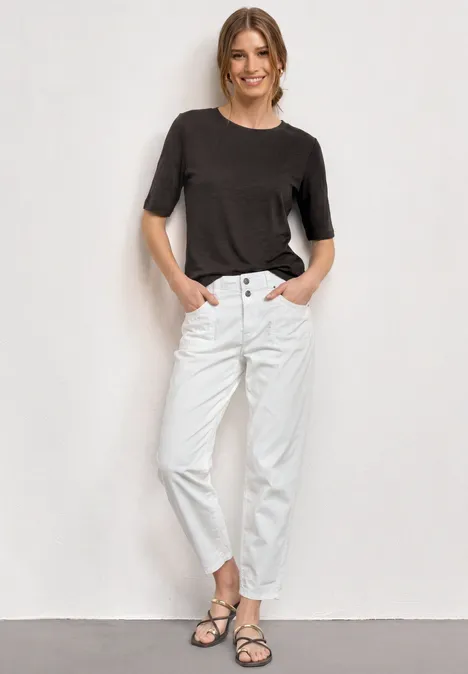 Basic Shirt met ronde hals in Unifarbe black coffee Persoon staat voor een witte muur, draagt een donker shirt met korte mouwen, witte broek en sandalen.
