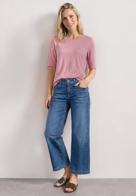 Model trägt rosa T-Shirt, hochgeschnittene blaue weite Jeans und schwarze Sandalen vor neutralem Hintergrund.