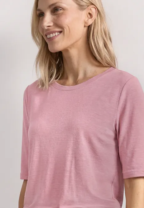 Eine Frau mit blondem Haar trägt ein schlichtes rosa T-Shirt vor hellem Hintergrund.