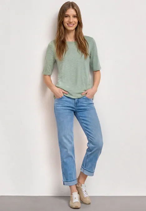 Basic Shirt met ronde hals in Unifarbe eucalyptus green Vrouw in een blauwe jeans en los groen shirt staat ontspannen voor een lichte muur.