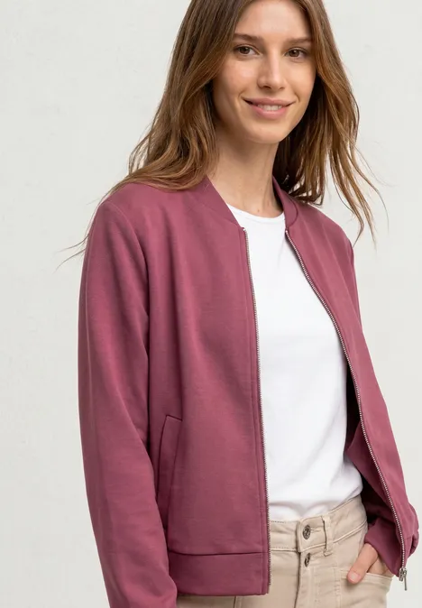 Femme souriante en bomber bordeaux, t-shirt blanc et pantalon beige, mains dans les poches.
