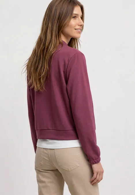 Femme en pantalon beige et t-shirt blanc, portant un sweat bordeaux, vue de profil tournant la tête en arrière.