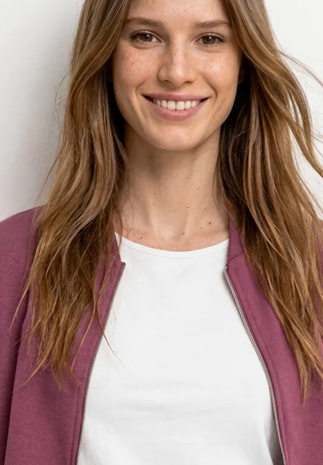 Une femme souriante aux longs cheveux bruns porte un t-shirt blanc et une veste rose.
