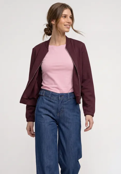 Eine junge Frau in burgunderroter Jacke, rosa Shirt und blauer Jeans lächelt, Haar im lockeren Dutt.