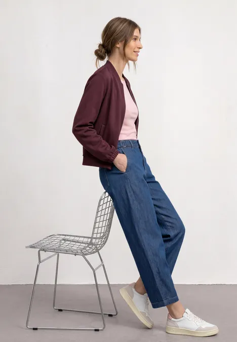 Junge Frau lehnt seitlich an Drahtstuhl, trägt bordeauxfarbene Jacke, rosa Shirt, weite Jeans und weiße Sneaker.