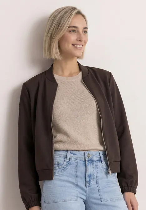 Frau mit blondem Bob trägt braune Bomberjacke, beigen Pullover und blaue Jeans vor neutralem Hintergrund.
