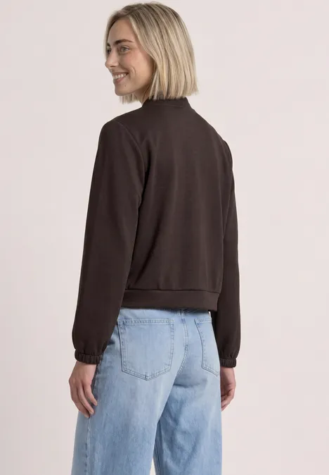 Frau in braunem Pullover und Jeans blickt lächelnd über die Schulter.