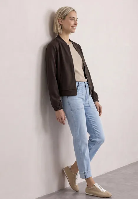 Junge Frau mit schwarzer Jacke, beigem Shirt und blauen Jeans lehnt lässig an heller Wand.