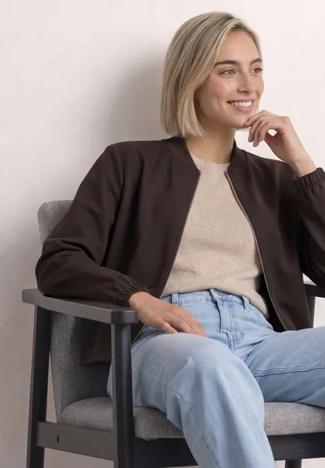 Eine junge blonde Frau sitzt lächelnd auf einem grauen Sessel, trägt eine beige Bluse, braune Jacke und blaue Jeans.
