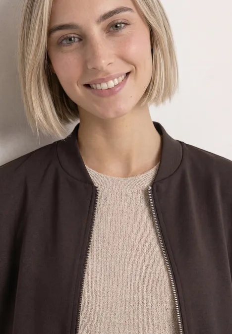 Eine Frau mit kinnlangem blonden Bob, trägt eine Jacke über einem hellen Pullover und lächelt.