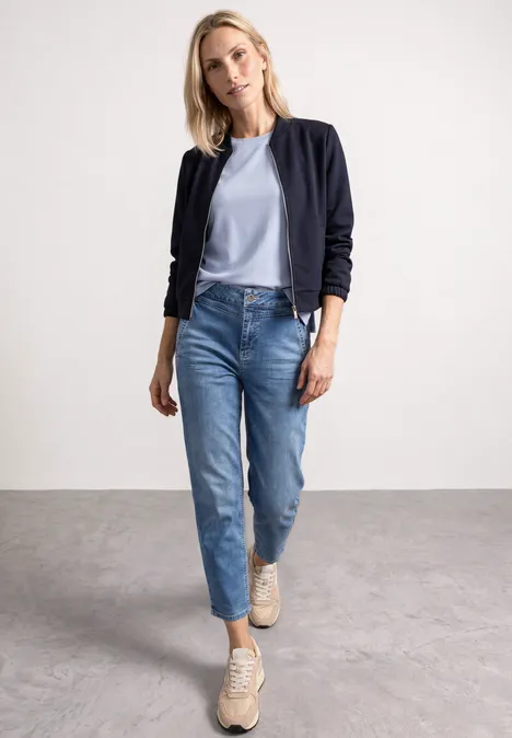 Frau in Bomberjacke, hellblauer Bluse, Jeans und Sneakern geht selbstbewusst in schlichtem Raum.